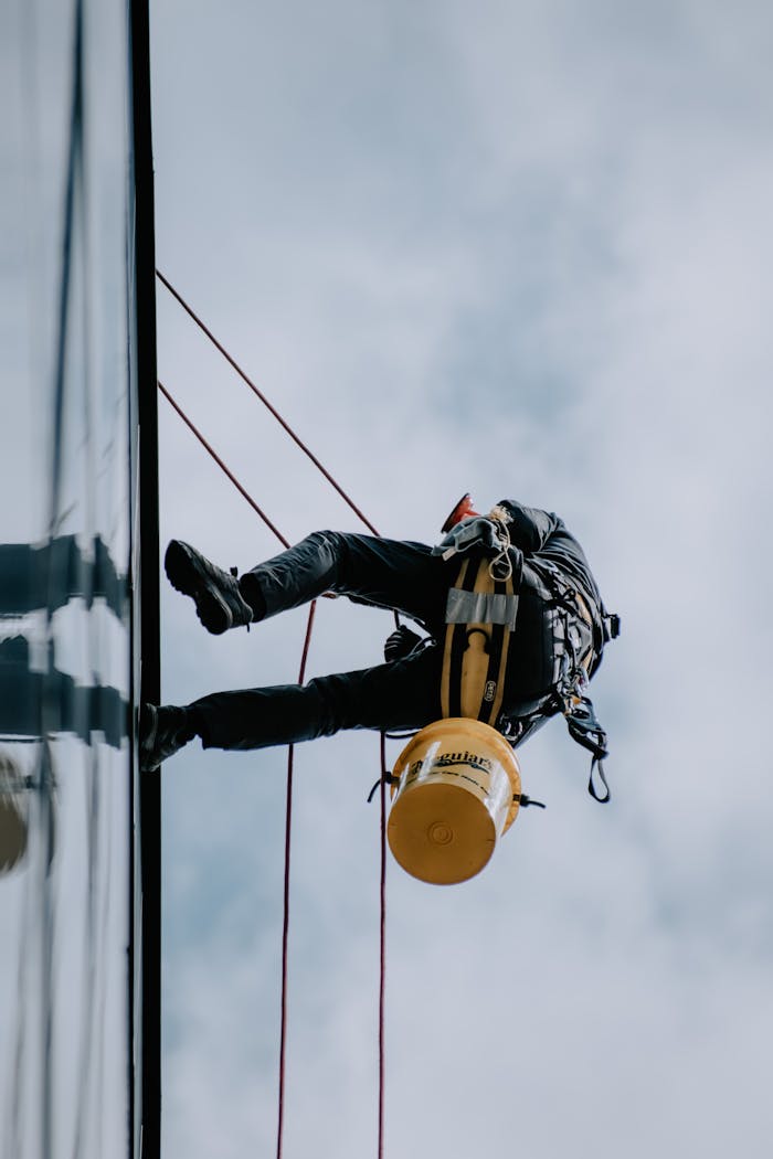 gallery-6 High-angle view of a window washer scaling a building facade with cleaning gear and ropes.