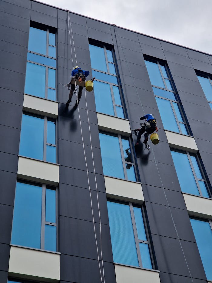 hero-img-01 Two workers clean a modern building facade at height with safety ropes.
